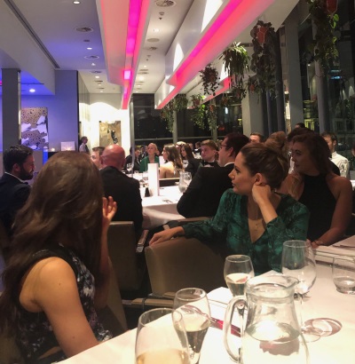 Three young women in the foreground at a table with wineglasses, turned to look across a room with other formally dressed people seated round set tables